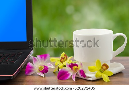 White coffee cup with hot coffee and steam and balck laptop computer with blue screen and freh yellow and pink orchid on wooden table in nature bokeh background