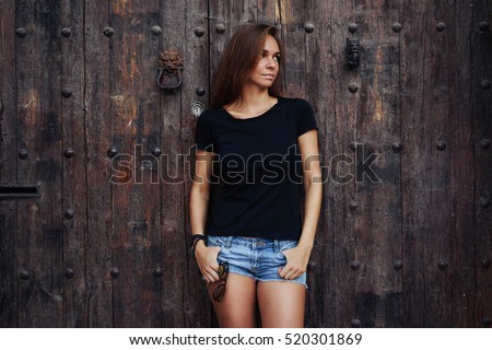 A street photo of a young beautiful female wearing black blank t-shirt and blue jeans shorts standing on the wooden door background. Empty space for text or design.