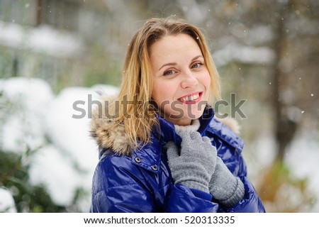 Portrait of charming woman against the background a winter landscape. She is dressed in bright blue down-padded coat, without headdress. Snowflakes on her hair. Woman has a fine smile and sad eyes.