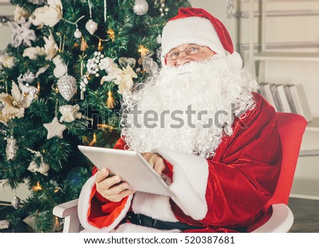 Portrait of happy Santa Claus holding Christmas letter using tablet on the armchair at home in the living room