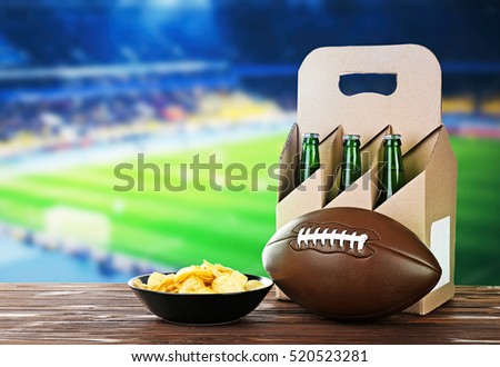 Beer with snack and ball on wooden table against football field background. Sport and entertainment concept.