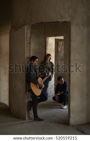 Full length shot of a young musician with an acoustic guitar leaning by the wall while rock band members standing in the background. 
