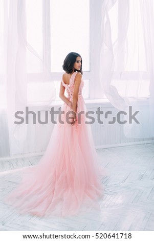 brunette girl standing in a bright room. She is dressed in luxurious, fluffy, pink dress. Gentle puppet's face. The European image of the bride. Fantasy Photo high key style. curtains flutter