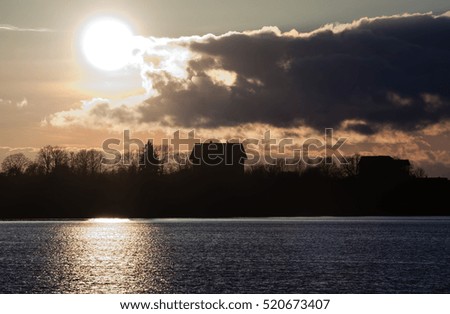 sunset in autumn, bright yellow, brown, dark blue and black, round the sun above the ground, at the bottom the black house and trees, patches light on water, solar path pond, heavy gray clouds,
