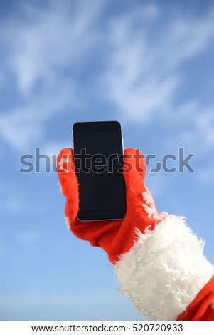 Happy Christmas with busy Santa Claus using touch screen smart phone. Close up image of telecommunication technology on festive blue sky outdoors background