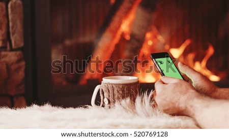 Unrecognizable Man Hands Using SmartPhone by the Burning Fireplace - Close Up. Male hands with phone green screen and cup by cozy fireside. Searching internet, using app.