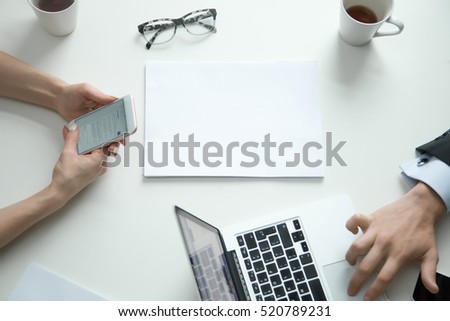 Top view of a white office desk, female hands with phone, male texting on the laptop, modern workplace, blank paper with copy space for text. Business concept photo, close up