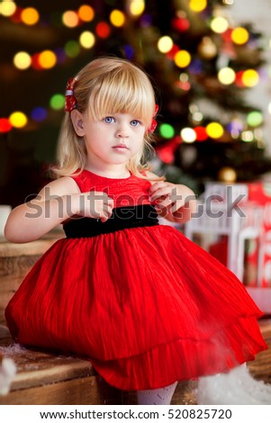 Adorable little blonde girl  in a red dress sitting on a floor near christmas tree with a boke background. New year photo.