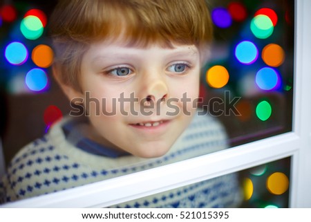 Adorable little blond kid boy near window and looking outdoors on Christmas time, indoors. Happy child with lights on background. Kid waiting for gifts and Xmas miracle.