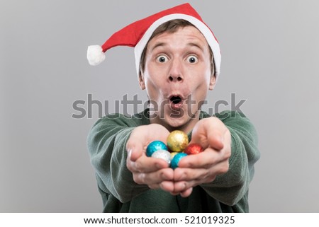 Amazed young man in christmas red hat holding multicolored decoration balls for christmas tree on grey background