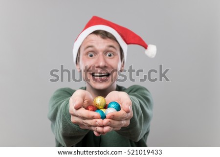 Amazed young man in christmas red hat holding multicolored decoration balls for christmas tree on grey background
