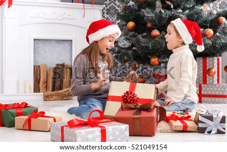 Cute happy excited children, boy and girl in santa hats unwrap christmas present box on holiday morning in beautiful room. Sister and brother open Xmas gifts near decorated tree and fireplace.