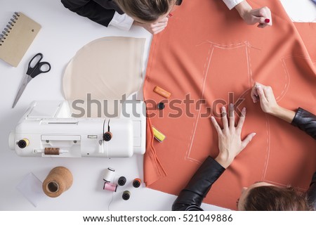 Top view of two girls making a pattern on a red piece of material. Sewing machine is standing on the table beside them. Royalty-Free Stock Photo #521049886