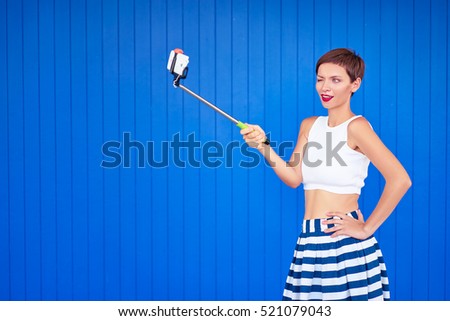 Colorful portrait of pretty young woman with haircut taking selfie on smartphone. Blue background.