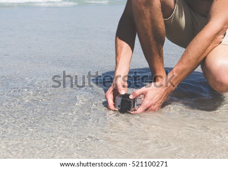 small modern action camera in waterproof housing filming in shallow water on beach, man holding recording equipment sitting outdoors 
