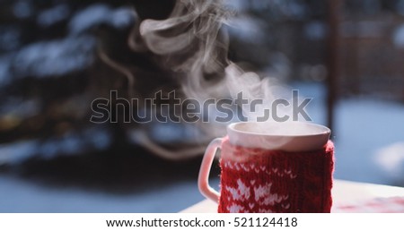 Steaming Cup of Hot Coffee or Tea standing on the Outdoor Table in Snowy Winter Morning. Cozy Festive Red Mug in knitted wear with a Warm Drink in Winter Garden. Christmas Morning Concept