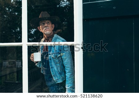 Farmer with beard and hat holding cup of coffee looking out window of old farm.