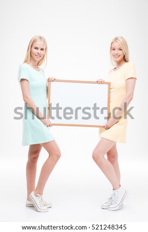 Picture of two ladies holding white blank desk. Isolated over white background. Looking at the camera.