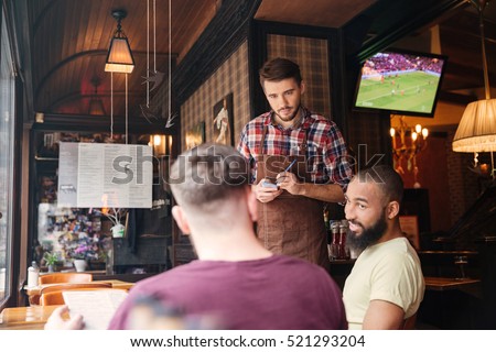 Handsome young waiter in brown apron taking an order from two relaxed men in bar  Royalty-Free Stock Photo #521293204