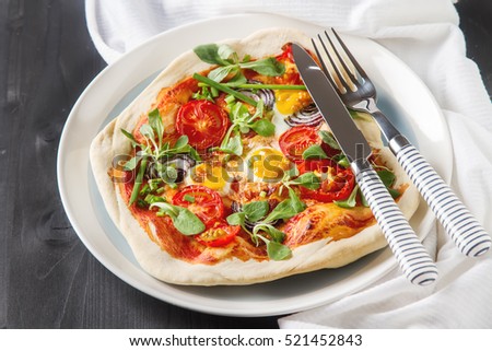 Home Italian pizza with tomato and quail eggs salad on a white plate, fork and knife striped. Black wooden table
