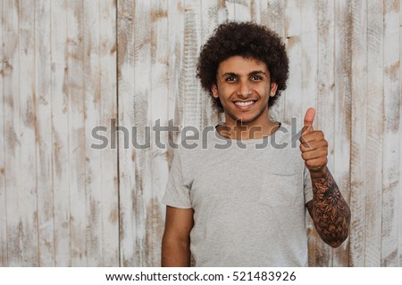 Do not worry be happy! Portrait cheerful curly haired  man  with perfect smile showing thumbs up. While standing against old wooden background