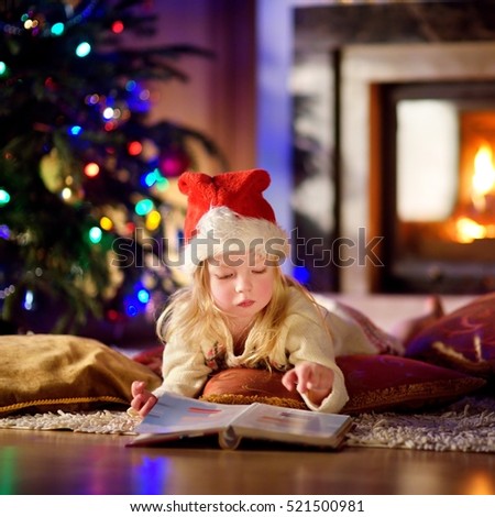 Adorable little girl reading a story book under a Christmas tree on Christmas eve at home