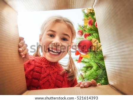 Merry Christmas and Happy Holidays! Cheerful cute child girl opening a Christmas present. Little kid having fun near Christmas tree indoors. View from inside of the box.