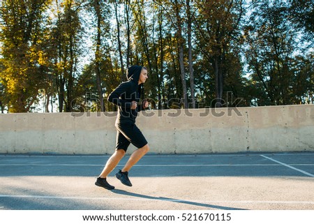 Young sportsman training on open-air stadium. Side view photo of running man in hood of sportswear, free space for text of advertisement.