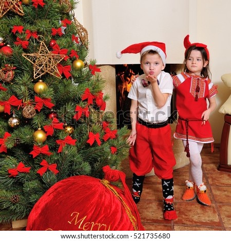 Good New Year spirit: Christmas tree, fireplace and gift bag - little cute girl with small red horns stand behind his blue-eyed cousin dressed in funny socks and elf costume sends an air kiss
