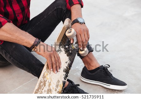 Cropped picture of dark skinned young man sitting near his skateboard. Against nature background.