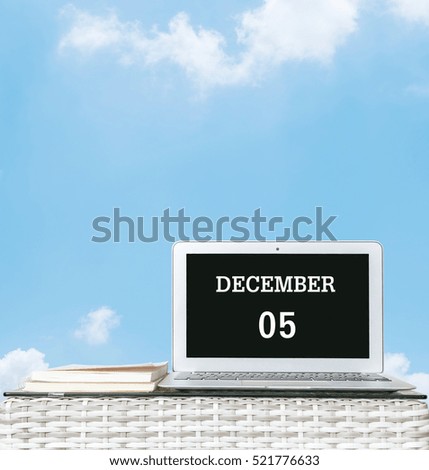 Closeup computer laptop with december 5 word on the center of screen in calendar concept on blurred wood weave table and book on blue sky with cloud textured background with copy space