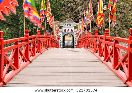 Red wooden Huc Bridge from the Ngoc Son Temple in Hanoi Vietnam leading to the exit door. Left and right waving flags in the wind Royalty-Free Stock Photo #521781394
