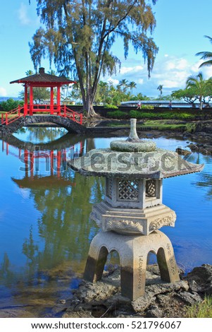 A traditional Japanese stone lantern sits at the edge of the pond in Liliuokalani Park, Hilo, Hawaii, with a bright red wooden bridge in the background.