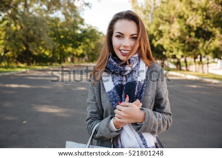 Picture of beautiful happy young woman wearing scarf smiling over nature background and holding bag. Looking at camera.