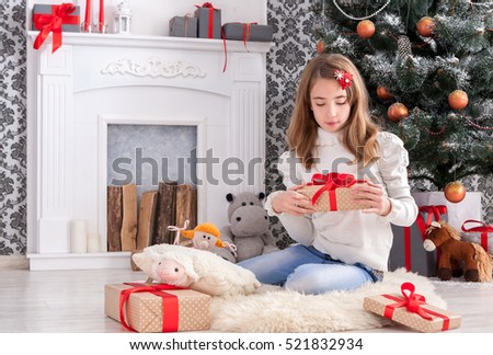 Beautiful happy girl unwrap christmas present box on holiday morning in beautiful room interior. Female child open Xmas gift near decorated fir tree and fireplace. Winter holidays concept