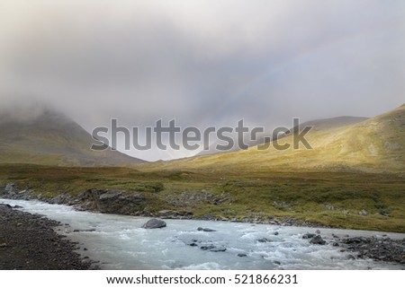 Stream of melted water flowing through tranquil landscape with rainbow between the mountain background