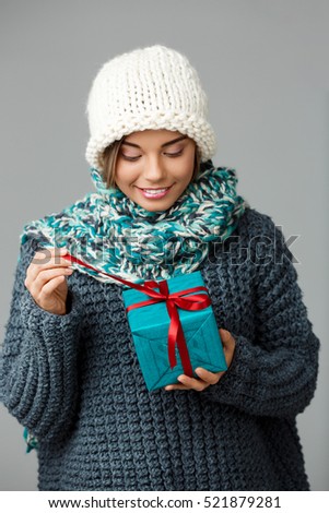 Young beautiful fair-haired girl in knited hat sweater and scarf smiling opening gift box over grey background. 