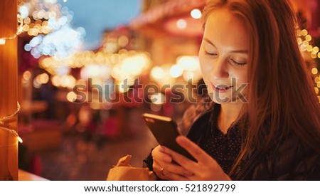 Woman Using Smartphone on European Christmas Market. Girl Enjoying Winter Holiday Season, Using phone, social networking, communicating, using app. Blurred Christmas Lights on background, dusk.