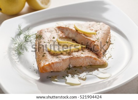 two baked in the oven salmon fillets with lemon slices seasoned with dill and pepper on a square white plate on a tablecloth

