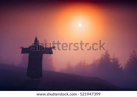 Fairy wizard in a black cassock standing on a hill and welcome raising sun above the foggy valley.