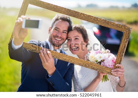 newlywed are going on honeymoon, they take a selfie through an empty frame, they have fun a lot. they are on a country road in the foreground and their car  is blur at the background. Shot with flare