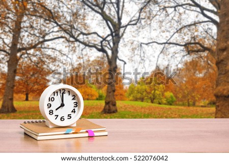 Clock and book on wooden table top on park in autumn season