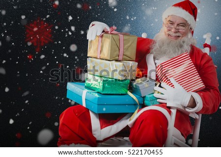 Portrait of cheerful Santa Claus holding Christmas presents on chair against snow with red flakes