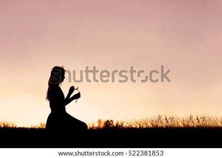 A silhouette of a peaceful young woman is sitting in a meadow in the country picking the petals off of a daisy wild flower at sunset.