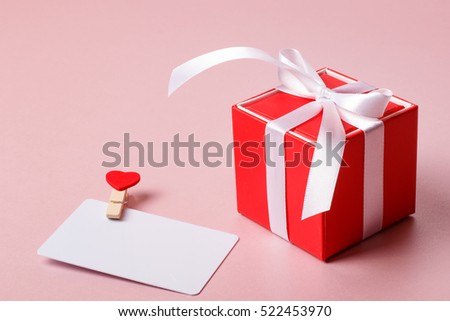 Valentine day composition: red gift box with bow and credit / visiting card template with clamp on light pink background.