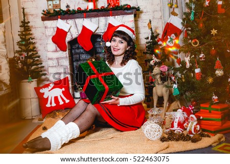 Beautiful brunette girl in santa hat sitting near fireplace, christmas decorated fir-tree and presents. Warm and cozy photo.Happy Holidays!