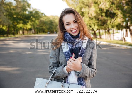 Photo of beautiful happy lady wearing scarf smiling over nature background and holding bag. Looking at camera.