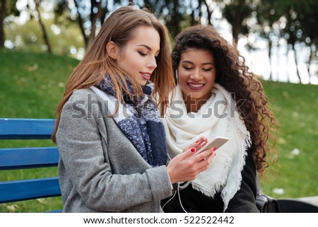 Photo of beautiful young women sitting on a bench and listening music together on earphones. Looking at phone. With grass on background.