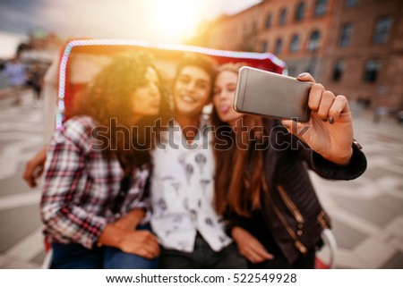 Young woman taking selfie on tricycle with friends. Young man and women riding on tricycle bike and taking self portrait. Focus on smart phone.