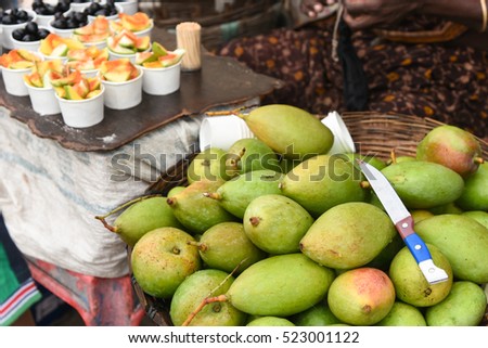 Heap raw green mango in a basket with knife and ripe black/purple sweet grapes and tangy mango salad with salt, chilli in paper cup arranged in fruit shop counter for sale,Chennai, Tamil Nadu, India.